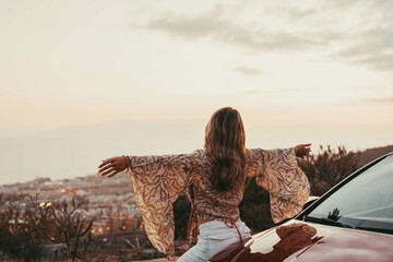 Wall Mural - Young beautiful woman enjoying summer and having fun sitting on the hood of the car opening arms feeling good and free. Freedom concept and happy lifestyle at sunset.