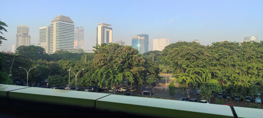 landscape of a parking lot in an urban area taken from the top of a building with lush and green trees with lots of parked cars and a misty skyscraper in the background during the day with a blue sky