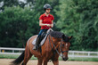 © qunica.com - A young girl is riding a horse at a ranch, wearing a red shirt and helmet for safety against a natural green backdrop.