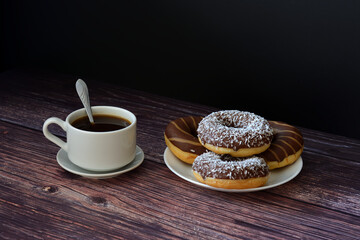 A cup of hot black coffee on a saucer with a spoon and a plate with four fresh donuts in chocolate icing on a wooden table.