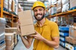 © AI Farm - Smiling warehouse worker in a yellow hard hat holding a cardboard box, standing in a large storage facility with shelves of goods.