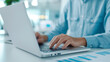 © TH_Stock - Close-up of a person typing on a laptop while analyzing financial reports. Focus on hands and laptop, with blue charts on the desk.