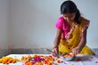 © Sourav Mittal - A young special needs woman wearing a pink and yellow dress is doing gudi padwa decoration in her home, sitting at a table near a white wall. She has some flowers at the bottom of her hand with a smal