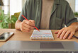 © Studio Romantic - Male college student writing to do list in his planner. Crop shot of young man sitting at table with paper notebook and laptop PC, holding pencil in hand and reading information about upcoming events