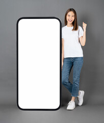 Wall Mural - Cheerful and beautiful Asian woman stands beside a large smartphone mockup with a blank screen against a grey background.