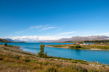 A scenic lakeside view with a charming stone church, surrounded by distant mountains and a nearby parking area. The bright blue sky and clear waters enhance the tranquil atmosphere, making it a pictu