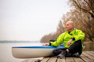 A middle-aged man sitting on the jetty and ready to go kayaking on the river