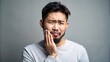 © Wanlop - Concerned young Asian man with a beard stands on a gray background clutching his painful lower jaw with both hands.
