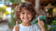 © Suralai - happy brunette boy child showing thumb in confirmation sign in a playground, wearing a white t-shirt,generative ai