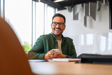 Wall Mural - confident project manager in glasses using smartphone and looking away while sitting at desk in offi