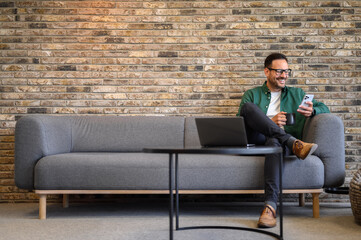 Wall Mural - handsome employee holding coffee cup and using social media apps over smart phone on sofa in office