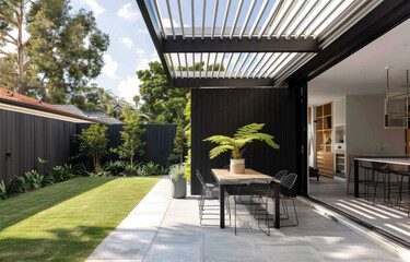 Modern backyard with black slat wall, dining table and chairs on concrete floor, white roof, green grass in background, trees, plants, and potted palm tree