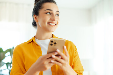 Close-up photo of a positive beautiful arabian or indian young woman in orange shirt holding smartphone in hands, typing message in social media, browsing internet, ordering products online in apps