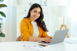 © Kateryna - Beautiful successful brunette arabian or indian young woman in an orange shirt, IT specialist, product manager sits at a workplace working in a laptop on a project, looking at camera, smiles friendly
