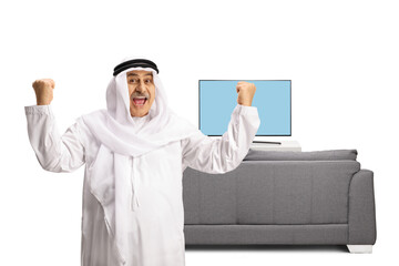 Wall Mural - Excited arab man with wearing dishdasha and gesturing happiness in front of tv and sofa