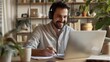 © inthasone - A smiling young man in headphones and glasses sits at a desk working on a laptop and making notes. Happy males in earphones watch webinars or training courses or computers and study online from home.