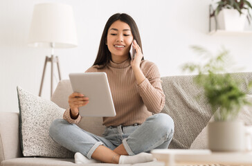 Wall Mural - Cheerful girl making order by phone using digital tablet, sitting on sofa at home