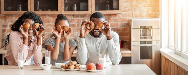 Funny african family having good time during breakfast at kitchen, making cupcake eyes, panorama