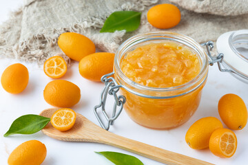 Wall Mural - Sweet kumquat jam or marmalade in glass jar and fresh raw whole and sliced fruits with leaves on white marble table background.