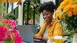 © Nataliia_Trushchenko - A woman is sitting at a table with a laptop and a cup of coffee. She is smiling and she is enjoying her time. The table is surrounded by potted plants, adding a touch of greenery to the scene