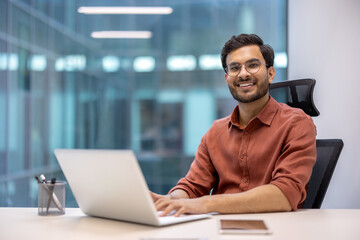 Confident young professional working on laptop in modern office. Smiling and looking at camera, embodies productivity and job satisfaction in a professional setting.