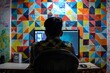© Inigo - Back view of a young man sitting at his desk in front of a computer with a colorful mosaic background