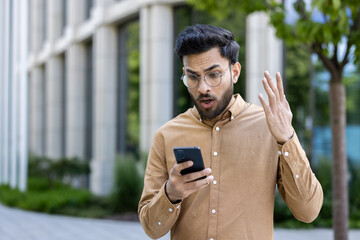 Man displaying shocked expression while looking at smartphone outside. Wearing glasses and brown shirt, reacting to unexpected news or message. Urban background includes modern building and greenery.