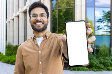 Smiling man with glasses holding blank phone screen outdoors near modern office building. Concept of technology, communication, and business. Ideal for promotional content or mockup design.