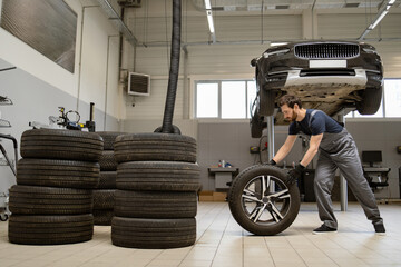 Mechanic in workshop handling car tire near vehicle lift. Organized garage space with multiple tire stacks. Professional auto repair environment showcasing maintenance and service.