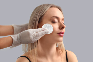 Wall Mural - Cosmetologist with cotton pad removing makeup from young woman's face on grey background