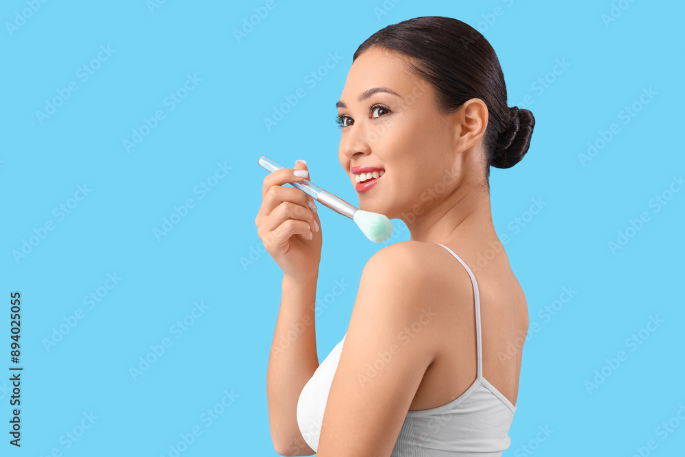 Young Asian woman with makeup brush on blue background, closeup