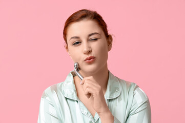 Sticker - Beautiful young woman in silk pajamas using facial massage tool on pink background