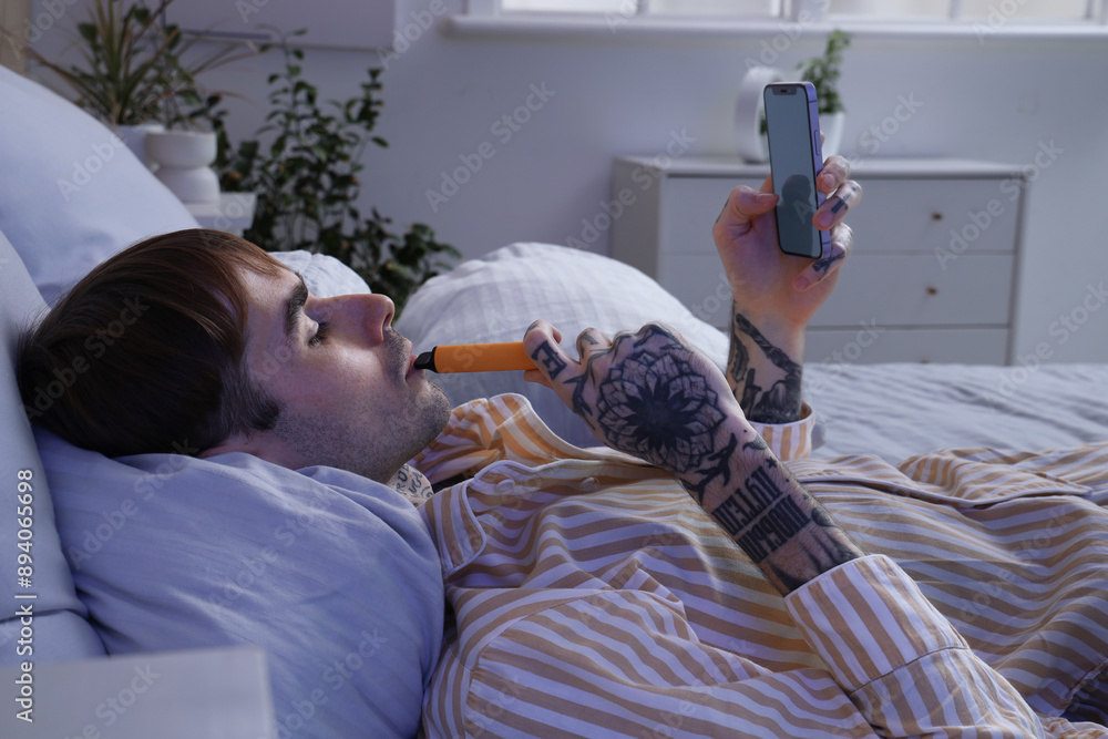 Young man in pajamas smoking disposable electronic cigarette and using mobile phone on bed at home
