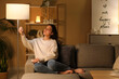 © Pixel-Shot - Young woman turning on lamp and sitting on cozy sofa in living room at night