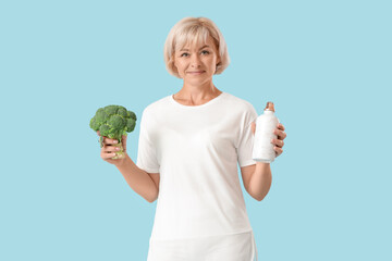 Poster - Sporty beautiful mature happy woman with bottle of water and fresh broccoli on blue background