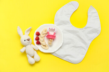 Wall Mural - Plate with baby nibbler, fruits, toy bunny and bib on yellow background