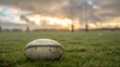 © narak0rn - A rugby ball resting on the grass, with goalposts and a few players warming up in the background under a cloudy sky