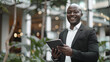 © Ruslan - Smiling African American businessman in a business suit with a tablet in his hands standing in a modern office