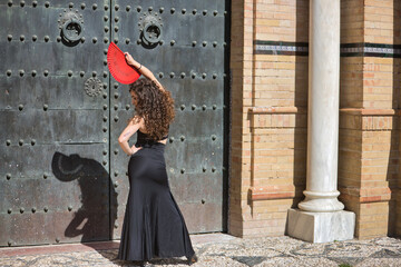 Canvas Print - Young, beautiful, brunette woman with black top and skirt, dancing flamenco with a red fan in front of an old, black metal door. Flamenco concept, dance, art, typical Spanish.