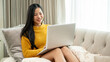 © bongkarn - An attractive Asian woman in comfy clothes is using a laptop computer on a couch at home.