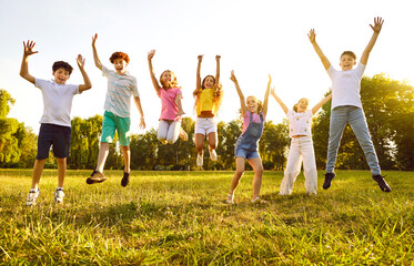 Wall Mural - Group of a smiling kids friends jumping with hands up on green grass in the park standing in a line. Happy children having fun together outdoors on a sunny summer day in casual clothes in the camp.