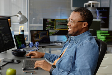 Side view shot of smiling male web developer of Black ethnicity in glasses sitting at working desk with multiple screens while using computer for code programming in office, copy space