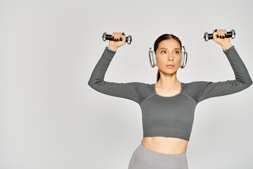 Wall Mural - Sporty young woman in active wear listening to music with headphones while holding two dumbbells on a grey background.