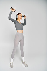 Wall Mural - A sporty young woman in active wear is exercising with dumbbells on a grey background.