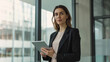 © VK Studio - Professional woman in a dark blazer holding a tablet, standing in a modern office with glass walls.