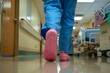 © Sourav Mittal - Close up of a nurse wearing blue scrubs and pink crocs walking in a hospital hallway, with a blurred background showing medical equipment, in a low angle shot from floor level, with the photo taken at