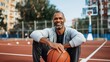 © Pinklife - A happy man sitting on an urban basketball court outdoors, holding a basketball and smiling, representing joy, fitness, and a love for sports in a city environment.