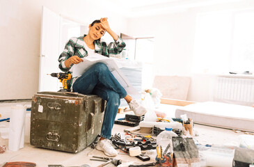 Young tired woman with house plan is sitting on box among building materials and inventory. Girl in casual clothes is thinking about home renovation.