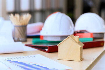 Small wooden house or home with engineering tools, blueprint and white safety helmet on messy table, real estate, property and construction project concept.