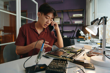 Repairman sitting at workplace with broken computer and discussing repair with customer on mobile phone